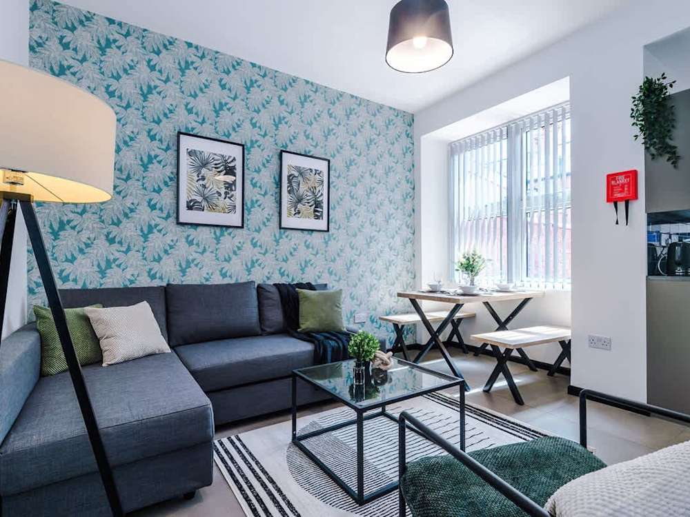 Living room of a Hostahome-managed house on Alpha Street, Salford, with a corner sofa, patterned feature wall and a dining table by the window.