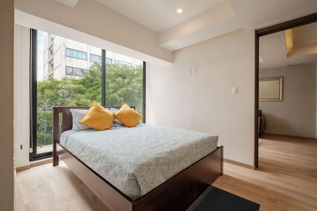 Restful bedroom with a comfy bed and golden accent pillows beside a floor-to-ceiling window, wake to city light near Reforma