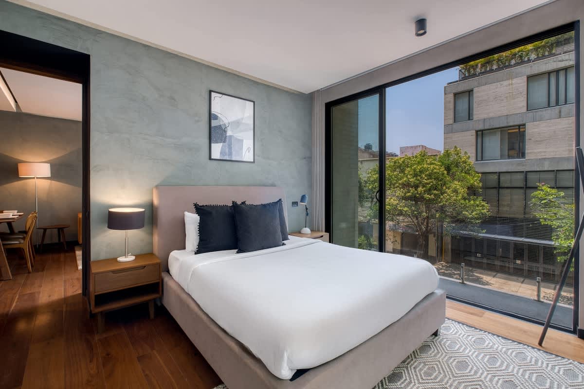 Restful bedroom with a plush king bed and a floor-to-ceiling window framing Roma Norte, wake to light and city views