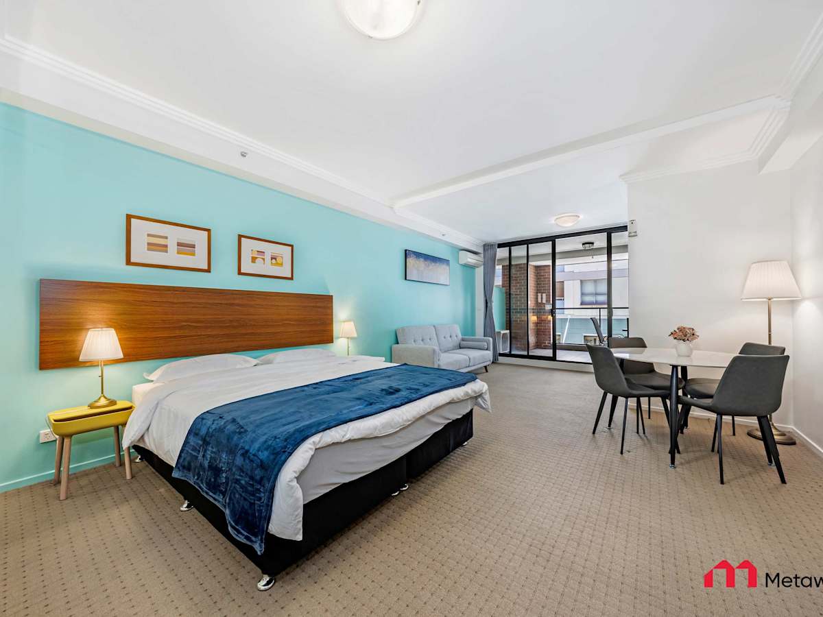 Spacious studio bedroom featuring a comfortable queen-sized bed, bedside lamps, teal feature wall, and a small dining table near the window.