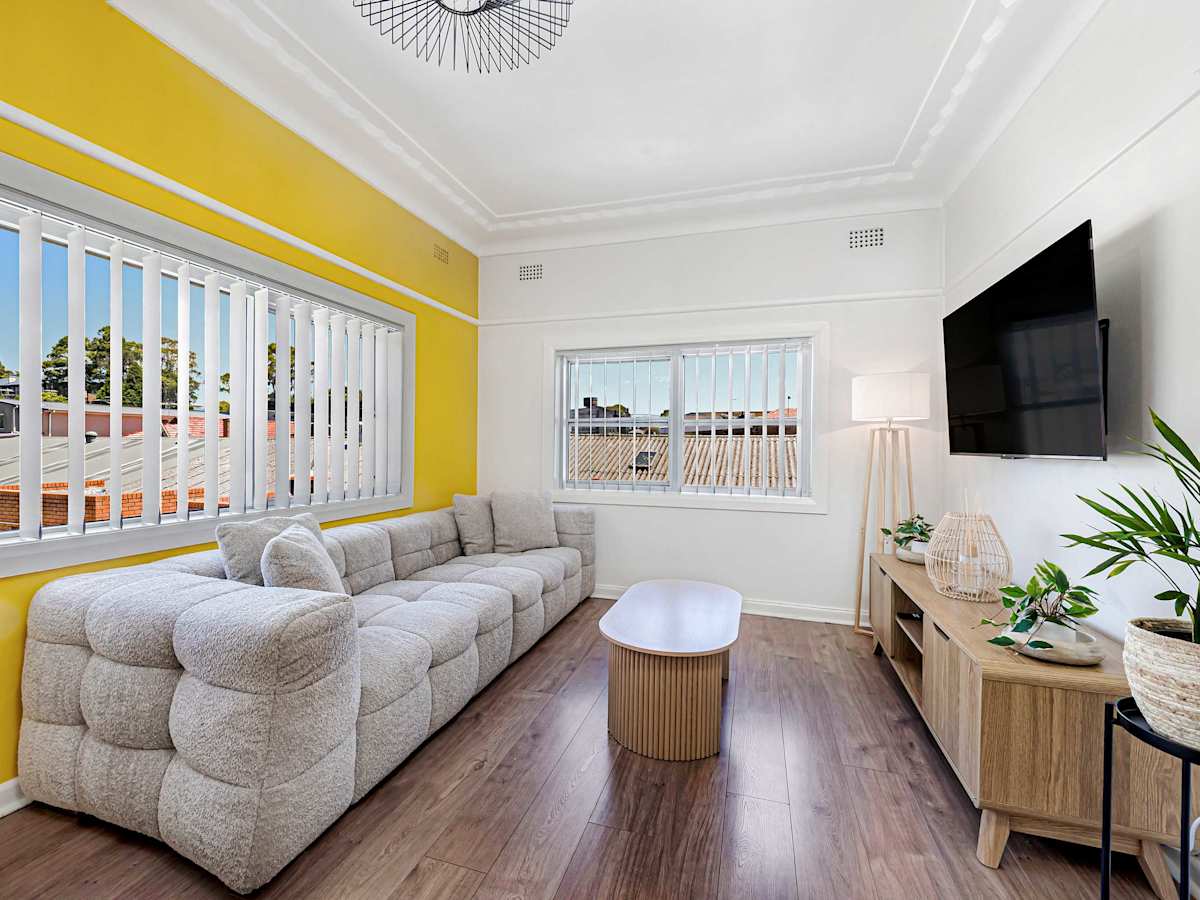 Living area with sofa, wall-mounted TV, and large windows with yellow feature wall.