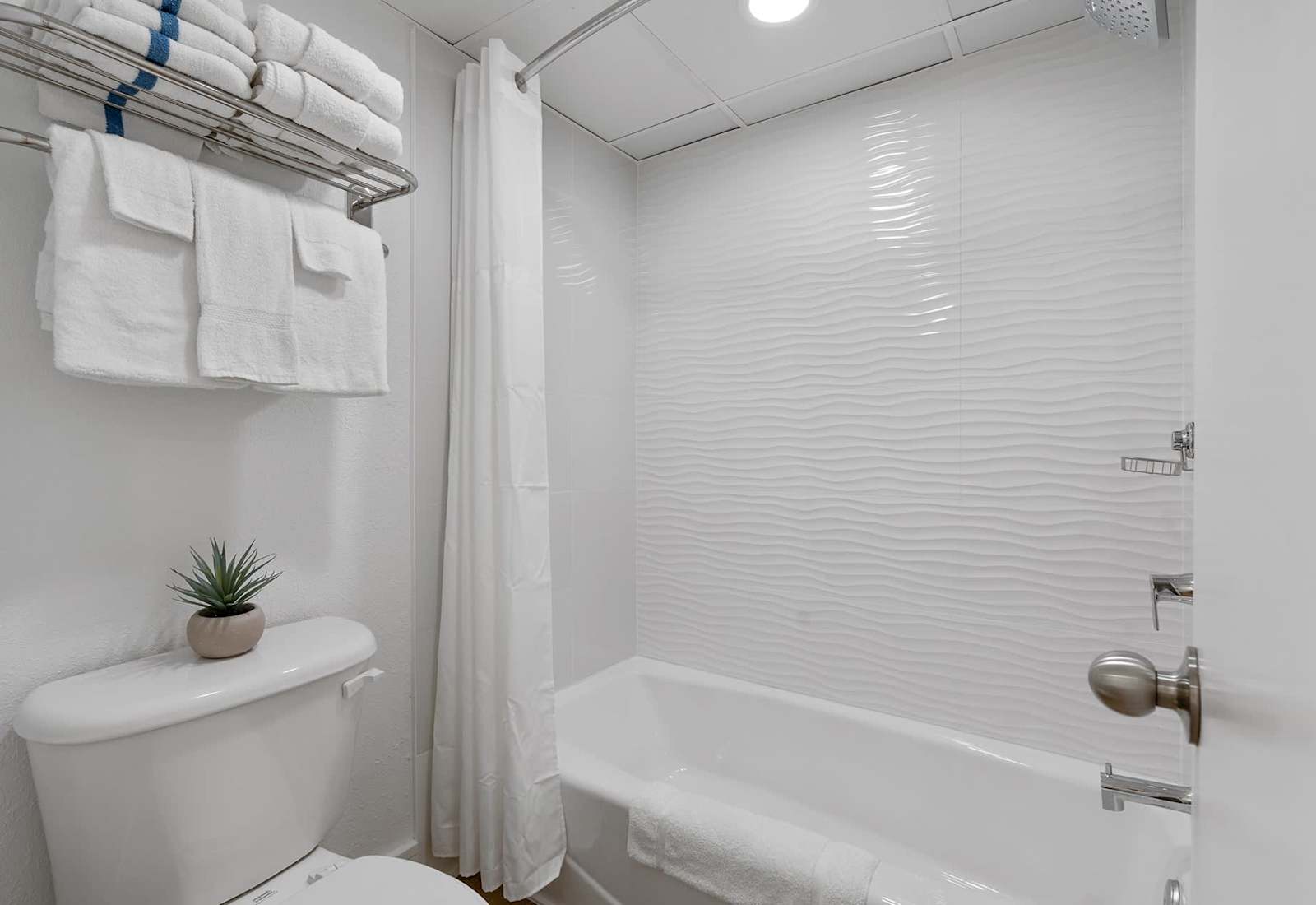 A bright, modern bathroom with crisp white tiles, a relaxing bathtub, and plush towels awaits your stay! 
