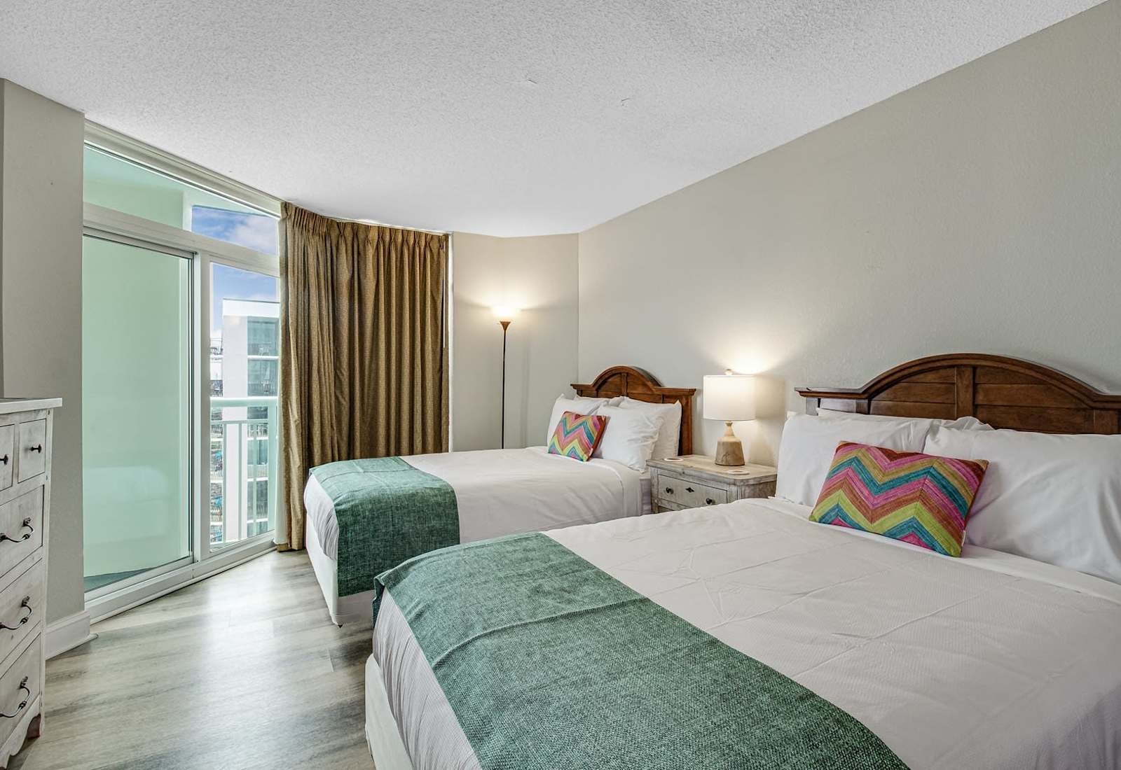 Spacious bedroom with two comfortable beds, soft coastal accents, bedside lighting, and sliding glass doors leading toward the balcony.
