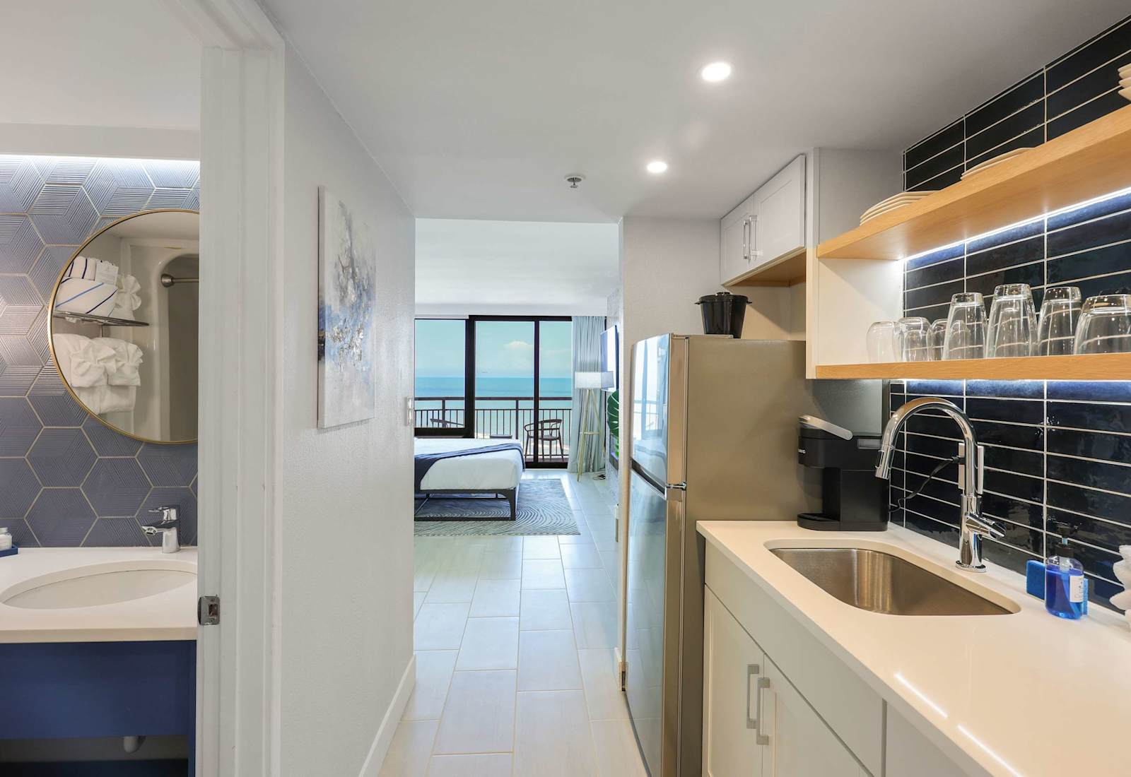 As you enter this elegant suite, you're greeted by a sleek kitchenette outfitted with glossy black tile, warm wood shelving, and a gleaming chrome faucet. Glassware and dinnerware are neatly arranged, ready for your morning coffee or evening toast.