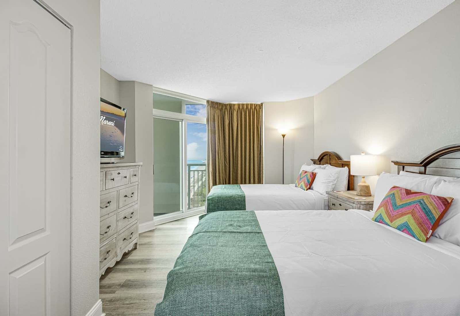 Comfortable bedroom with two beds, colorful coastal accents, TV, dresser storage, and balcony access with a peek of the ocean.