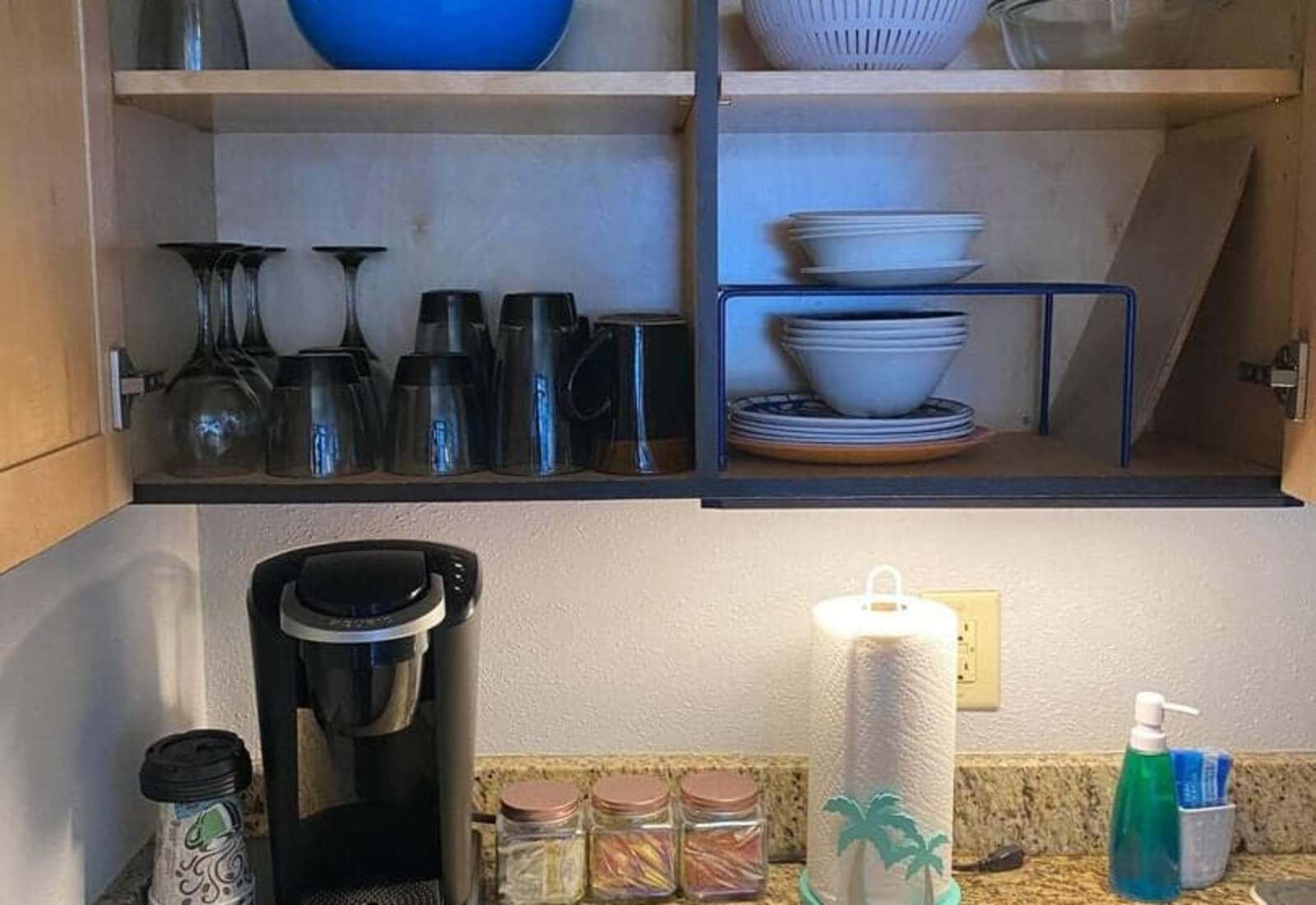 Well-stocked kitchen cabinet open above a granite countertop, displaying assorted dishes, bowls, and glassware alongside a coffee maker and paper towel roll.
