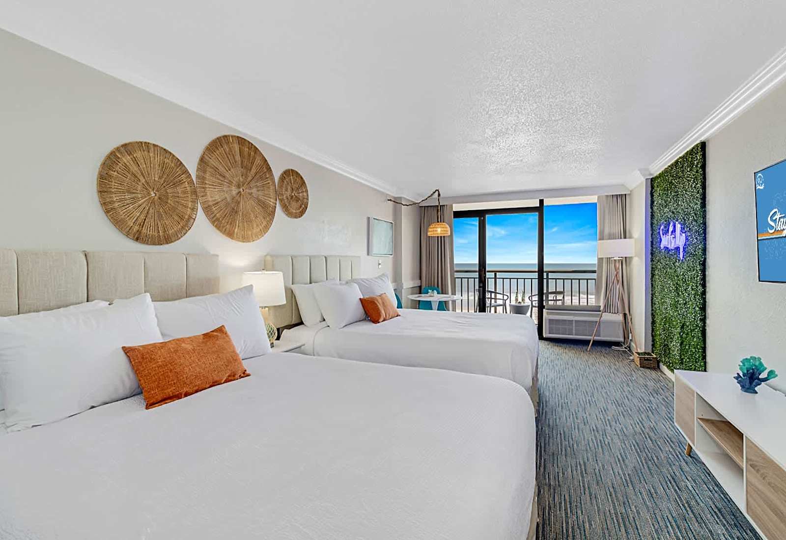 Bright, stylish oceanfront suite with two queen beds, a scenic balcony view, and modern decor. Features a lush accent wall with neon lighting, cozy seating area, and a large Smart TV. Perfect for a relaxing beach getaway!