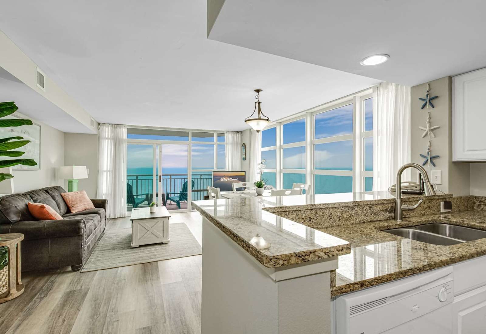 Open-concept kitchen and living area with granite countertops, bright coastal décor, and stunning ocean views from the windows and balcony.