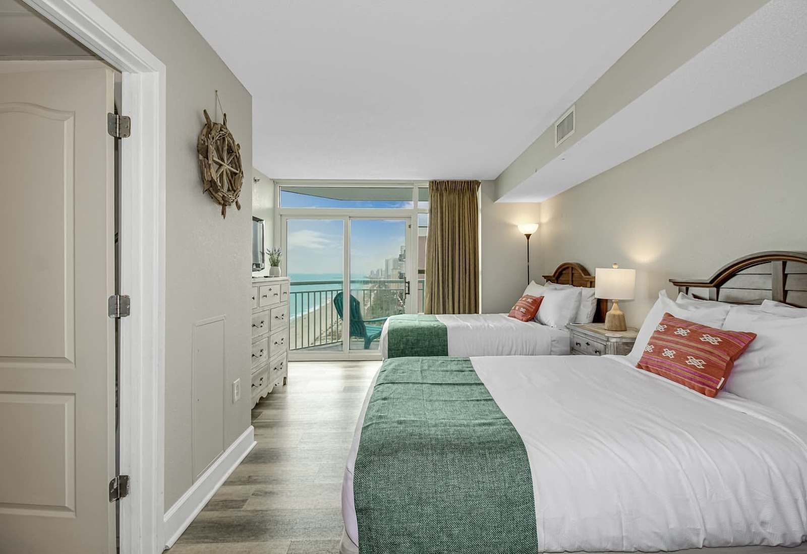 Rest easy in a bright double-bed room featuring crisp linens, colorful pillows, dresser storage, and natural light from the balcony doors.