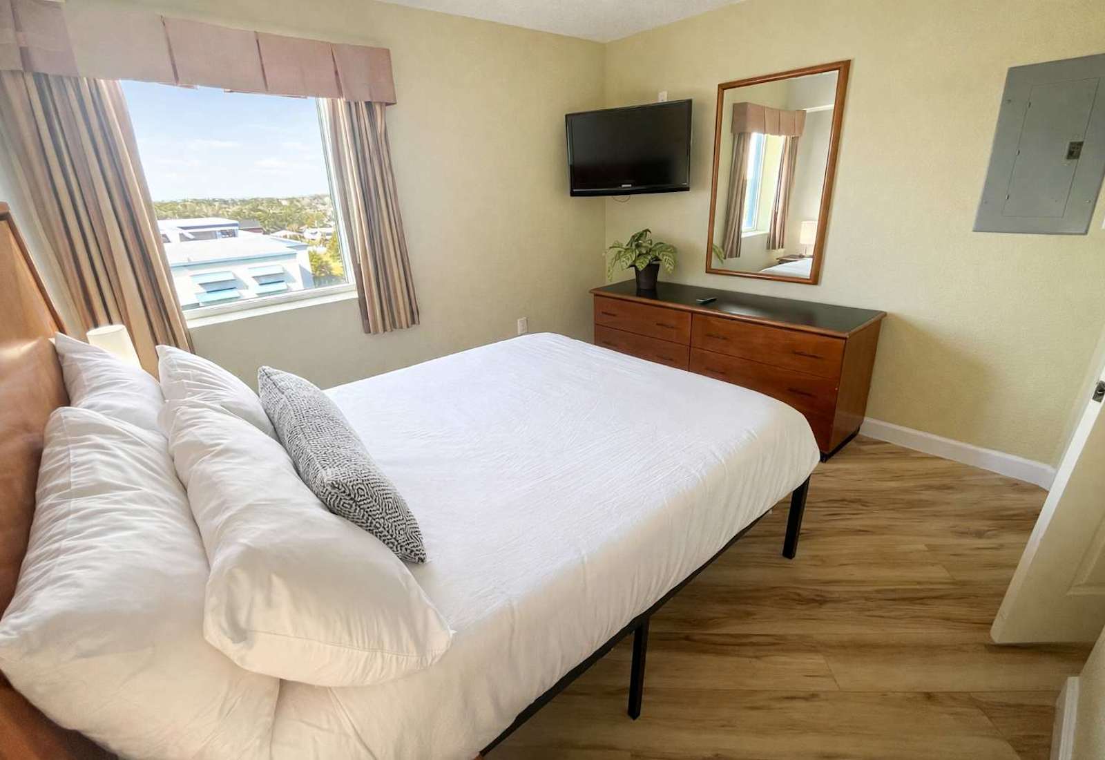 A cozy, sunlit bedroom featuring a crisp, neatly made bed with soft white linens and warm wood accents. Two modern bedside lamps add a gentle glow, while a large window brings in natural light. Simple, clean, and comfortable—perfect 