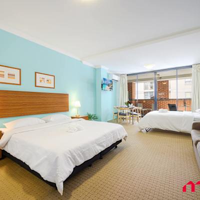 Spacious studio featuring a king-size bed near the entrance and a pull-up bed positioned near the balcony, with teal feature wall and bedside lighting.