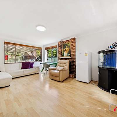 Spacious and light-filled living area featuring timber flooring, a white L-shaped sofa with accent cushions, and a beige recliner chair. The room includes a brick fireplace with artwork above, large windows with wooden blinds allowing natural light,