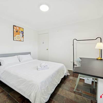 Comfortable bedroom featuring a queen-size bed, bedside table with lamp, study desk, and large window with blinds.