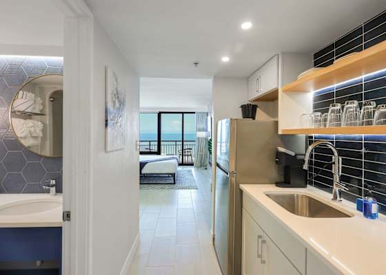 As you enter this elegant suite, you're greeted by a sleek kitchenette outfitted with glossy black tile, warm wood shelving, and a gleaming chrome faucet. Glassware and dinnerware are neatly arranged, ready for your morning coffee or evening toast.