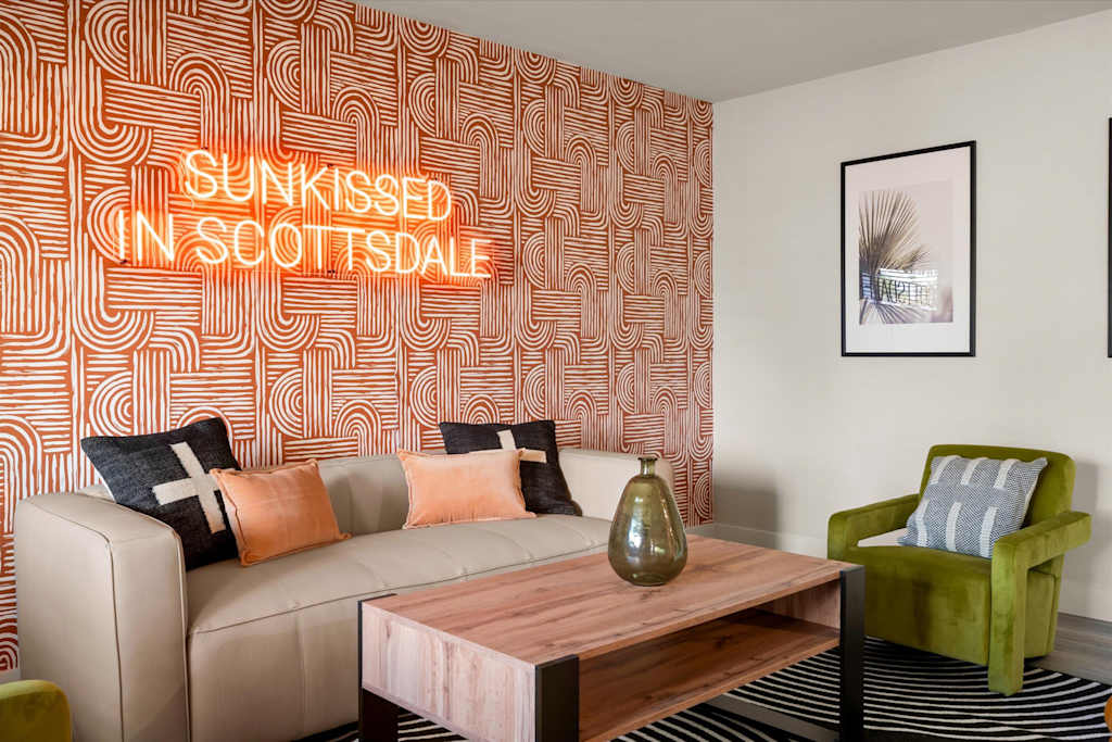 The Sunkiss — living area with a sofa, wooden coffee table, green armchair, and neon sign on patterned wallpaper