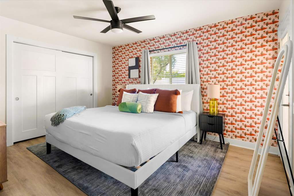 The Sunkiss — bedroom with a white bed, patterned accent wall, nightstand, window blinds, and standing floor mirror