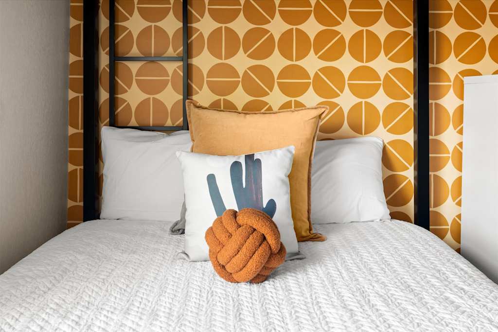 The Sunkiss — bed with white bedding, decorative knot pillow, and yellow geometric patterned wallpaper behind headboard