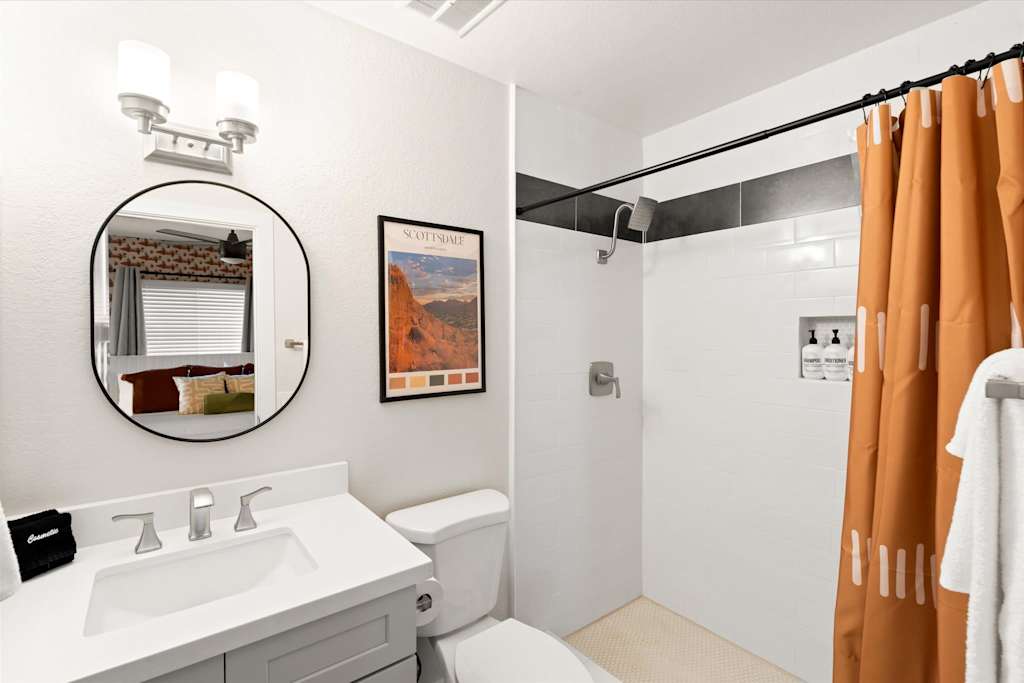The Sunkiss — bathroom with round vanity mirror, grey cabinets, white toilet, and shower with orange curtain