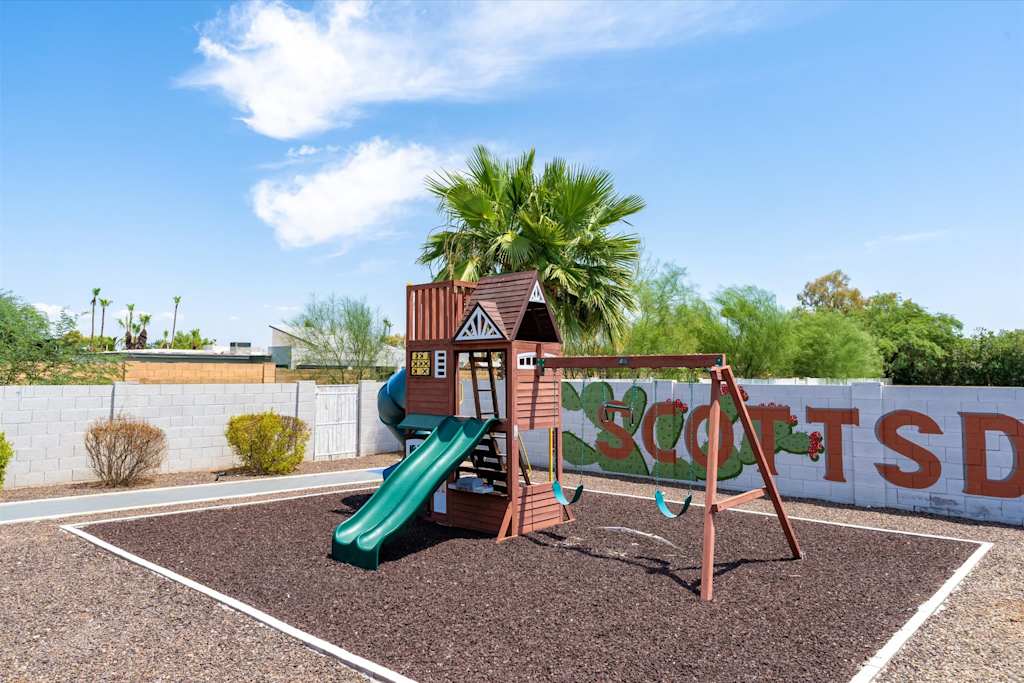 The Scottsdale Social — wooden children's playset with swings and a green slide situated on mulch in a fenced yard