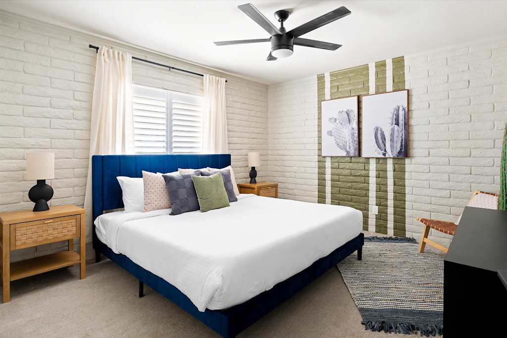 The Scottsdale Social — bedroom with a blue upholstered bed, white painted brick walls, cactus artwork, and ceiling fan