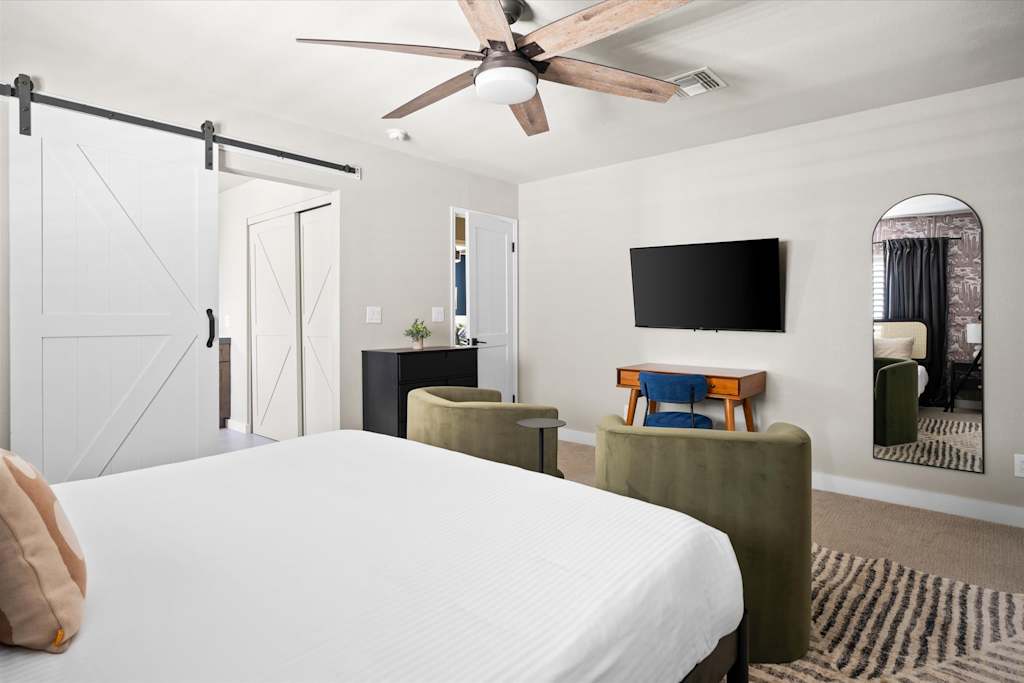 The Scottsdale Social — bedroom featuring a bed, two green velvet armchairs, desk, wall-mounted television, and sliding barn door