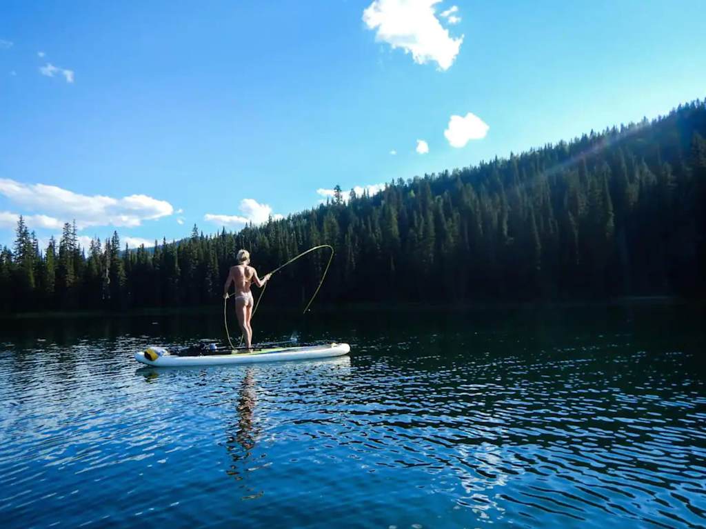 The Evergreen — person fly fishing from a stand-up paddleboard on a calm lake bordered by evergreen trees