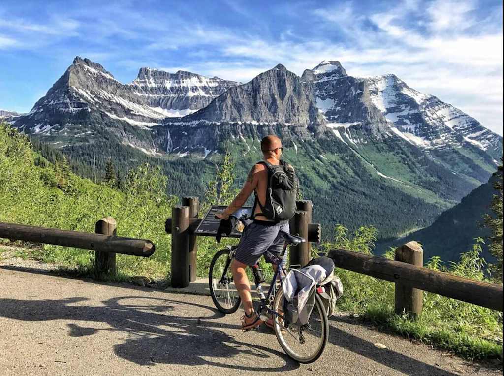 The Evergreen — cyclist with a bicycle paused at a roadside overlook facing snow-dusted mountain peaks