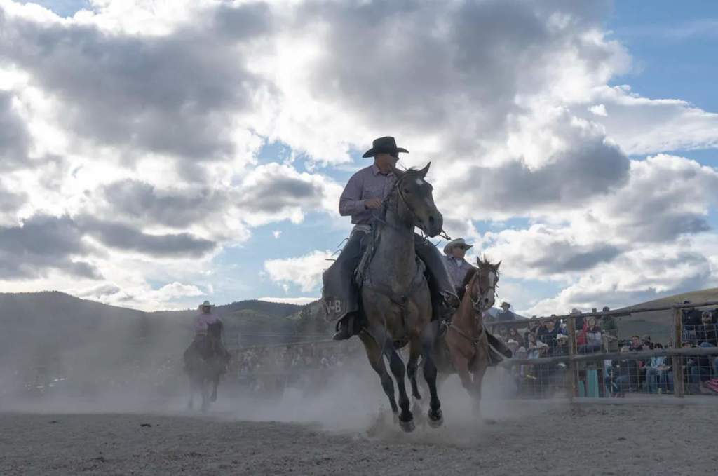 The Evergreen — cowboys on horseback galloping across a dusty outdoor arena under a cloudy sky