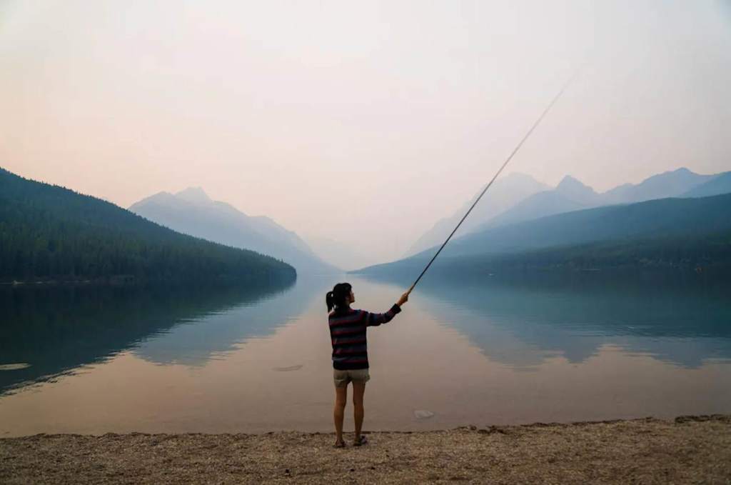 The Evergreen — person holding a fishing rod on the shore of a calm lake surrounded by mountains