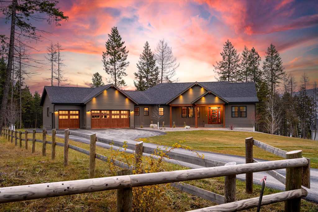 The Evergreen — front exterior of single-story house with attached garage, driveway, and split-rail fence at sunset