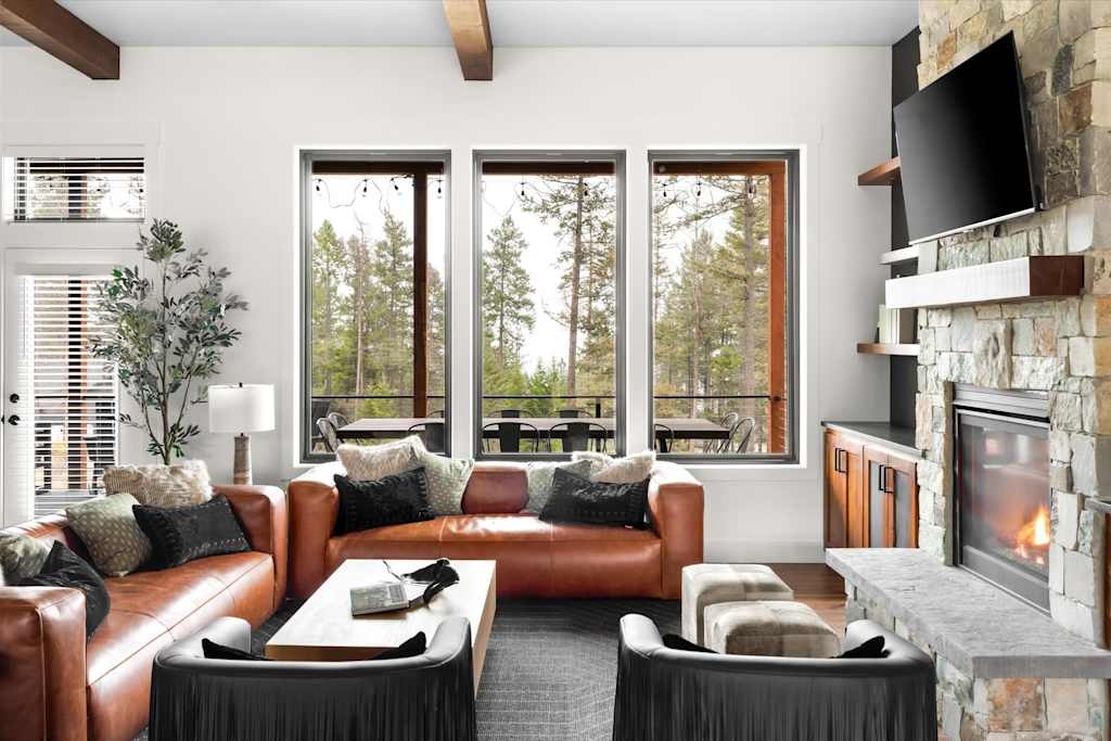 The Evergreen — living room with brown leather sofas, large windows looking out to trees, and stone fireplace