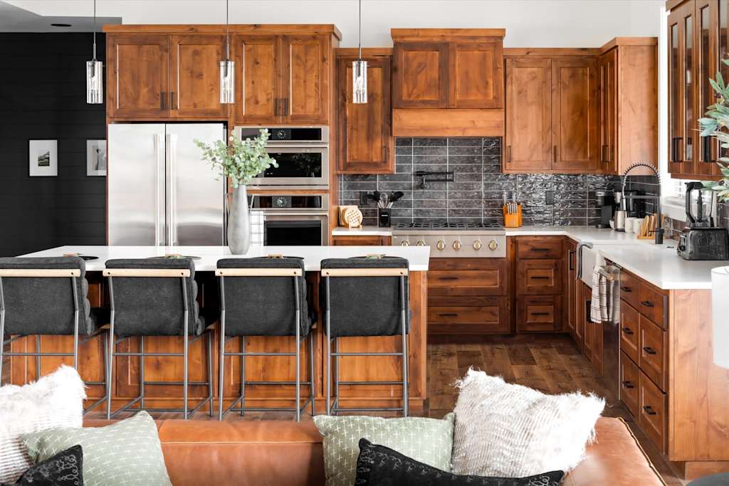 The Evergreen — kitchen with wooden cabinetry, stainless steel appliances, dark tile backsplash, and island bar seating
