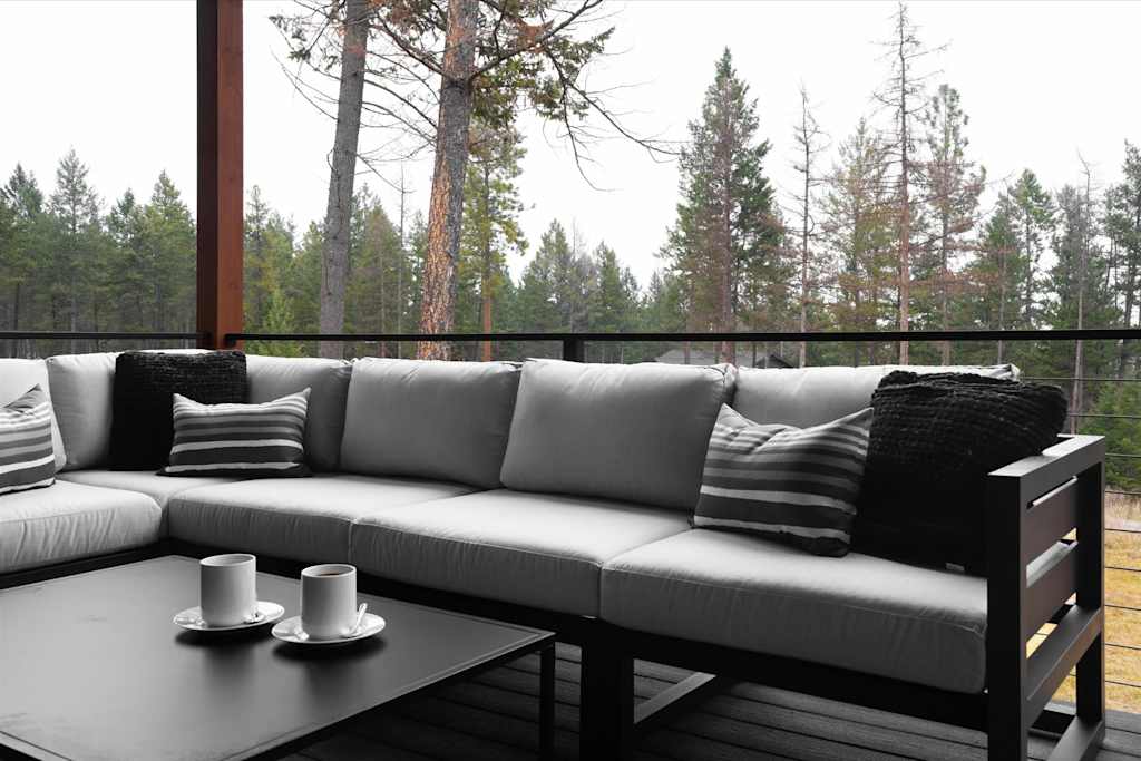 The Evergreen — covered outdoor deck with grey sectional sofa, coffee table with mugs, and surrounding pine forest