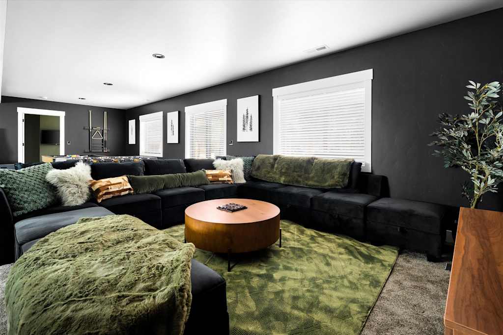 The Evergreen — basement living area with dark walls, large black sectional sofa, round coffee table, and green rug