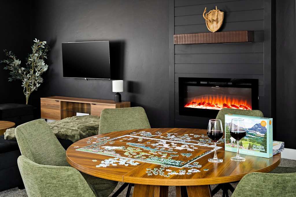 The Evergreen — round wooden table with puzzle and wine glasses, set in front of an electric fireplace