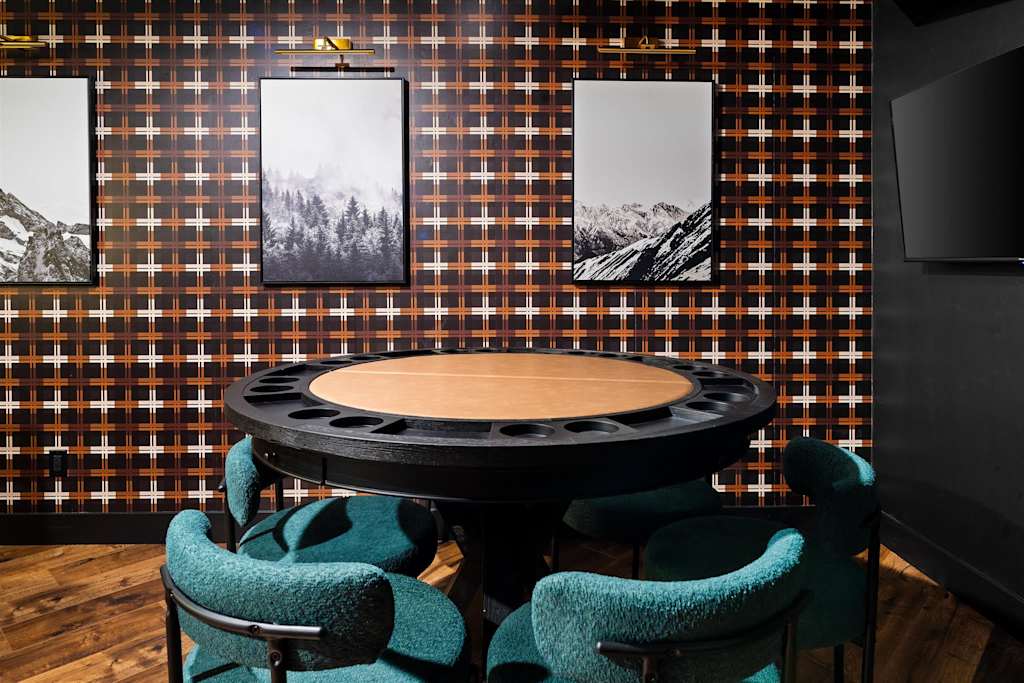 The Evergreen — round poker table surrounded by teal upholstered chairs in front of a plaid accent wall