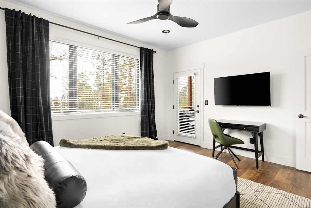 The Evergreen — bedroom with bed, wall-mounted tv, small work desk, and large window with blinds