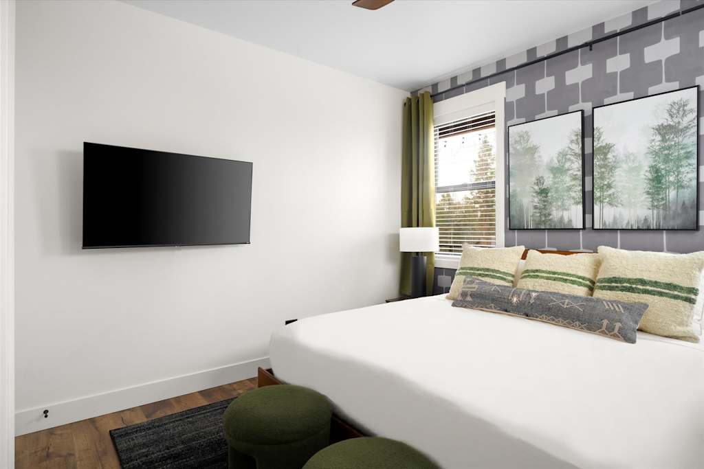 The Evergreen — bedroom with a wall-mounted flat-screen television, bed, two green ottomans, and a window