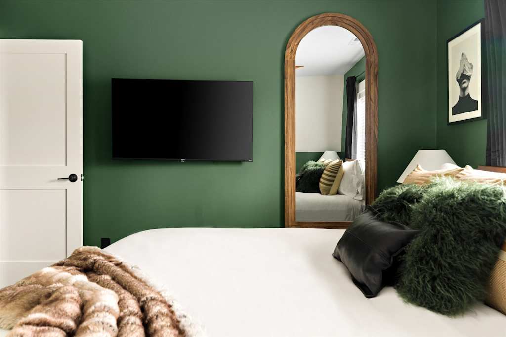 The Evergreen — bedroom with dark green walls, wall-mounted television, large arched mirror, and white bed
