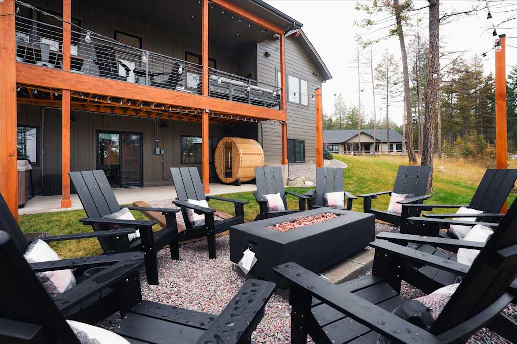 The Evergreen — outdoor patio area featuring black adirondack chairs around a fire pit, barrel sauna, and house exterior