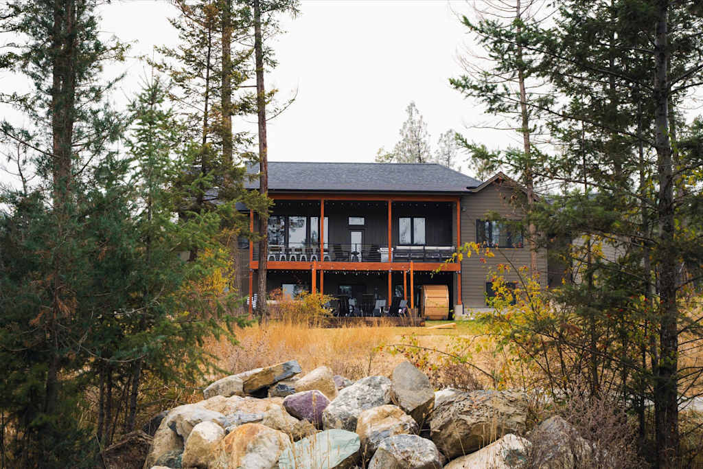 The Evergreen — rear exterior view of a two-story home framed by tall evergreen trees and rocks in foreground