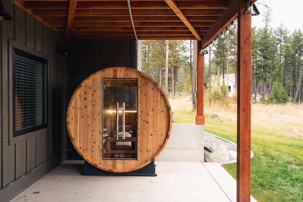 The Evergreen — wooden outdoor barrel sauna situated on a concrete patio beneath a covered porch