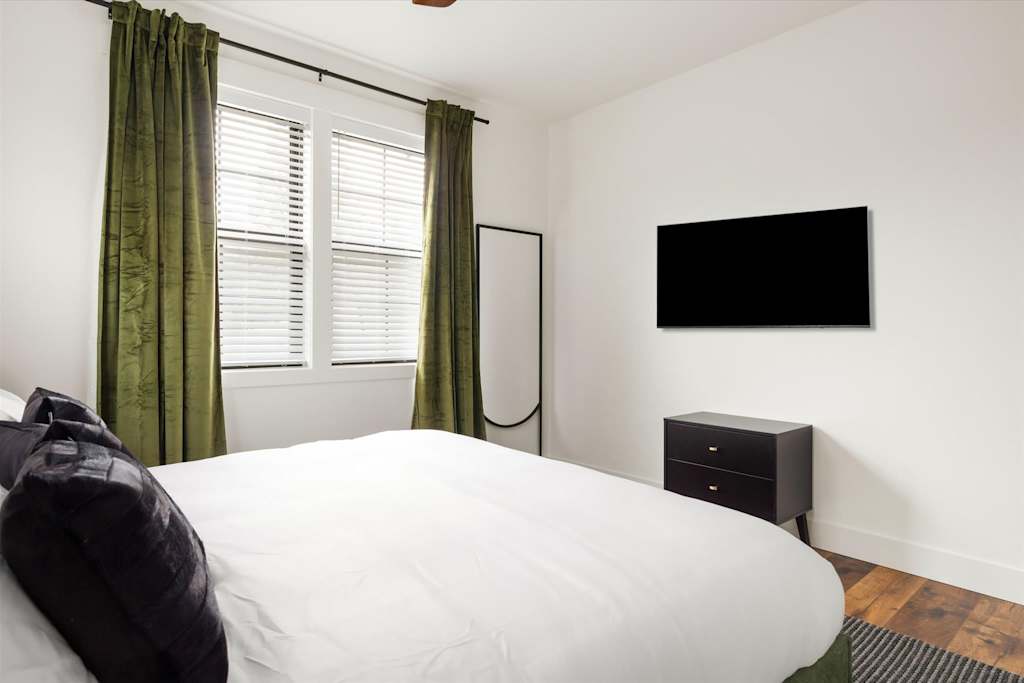 The Evergreen — bedroom view facing a wall-mounted flat-screen television above a black dresser next to windows
