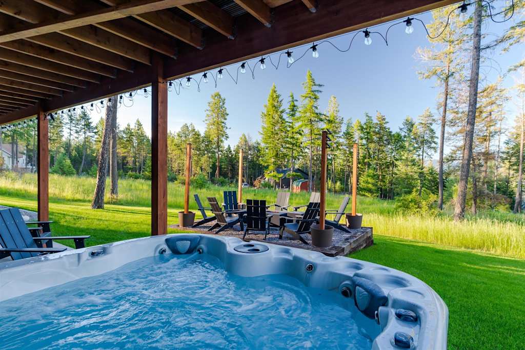 The Evergreen — outdoor hot tub on a covered patio overlooking a gravel fire pit area and woods