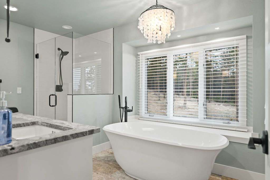 The Tamarack — white freestanding soaking tub beneath a window with blinds, beside a glass shower
