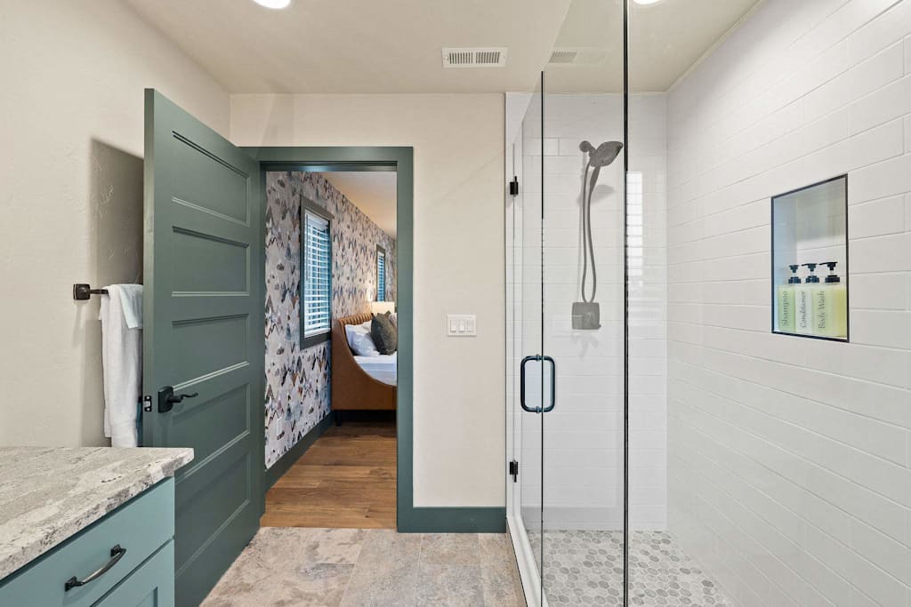 The Tamarack — bathroom with glass walk-in shower, green door opening to bedroom, and vanity counter in foreground
