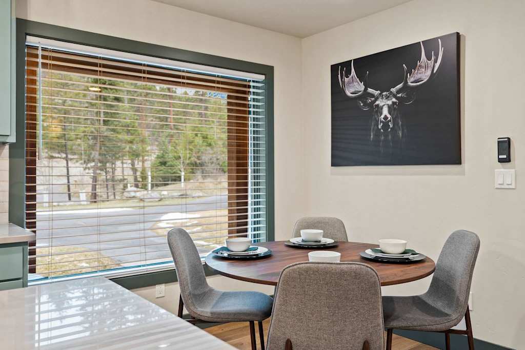 The Tamarack — dining area with round wooden table, four chairs, window blinds, and framed moose artwork