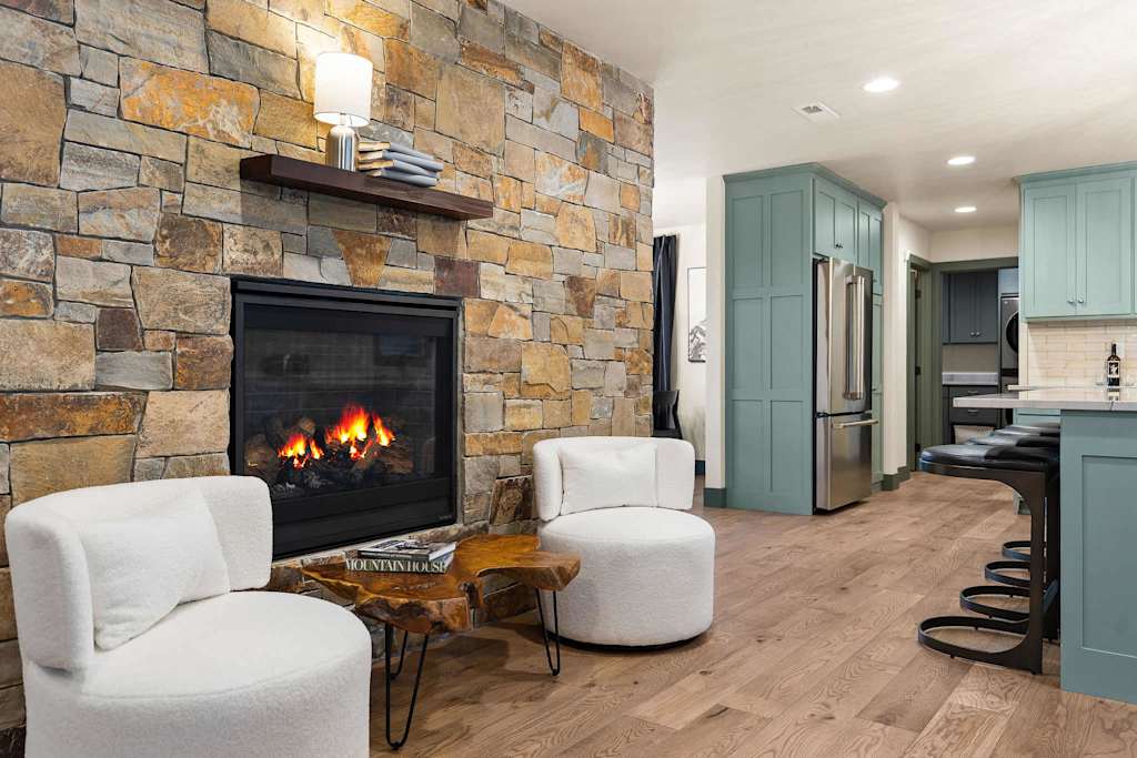The Tamarack — stone fireplace with lit fire, two white armchairs, live-edge coffee table, and kitchen beyond
