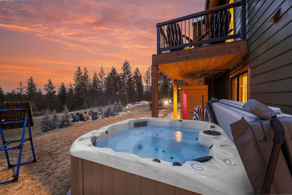 The Tamarack — outdoor hot tub on patio under upper deck at sunset with forest views