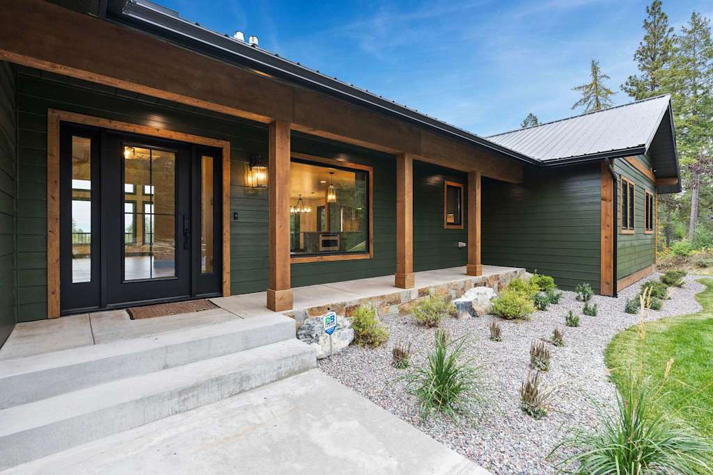 The Tamarack — front entrance porch of a dark green house featuring timber columns, concrete steps, and gravel landscaping